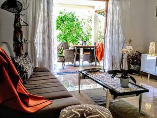 A cozy living room with a gray couch, orange throw, coffee table, and a potted plant. The room opens to a patio through sheer white curtains, featuring a dining set and greenery outside.