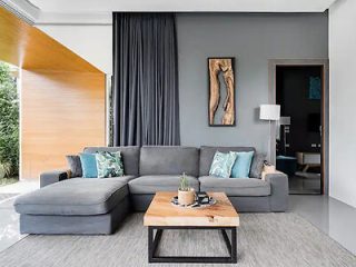A modern living room with a grey sectional sofa, wooden coffee table, and wall art. The room features large curtains, a floor lamp, and an open doorway leading to another room.