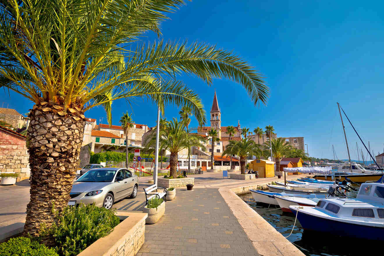 A waterfront scene with docked boats, palm trees, parked cars, historic buildings, and the bell tower of a church under a clear blue sky.