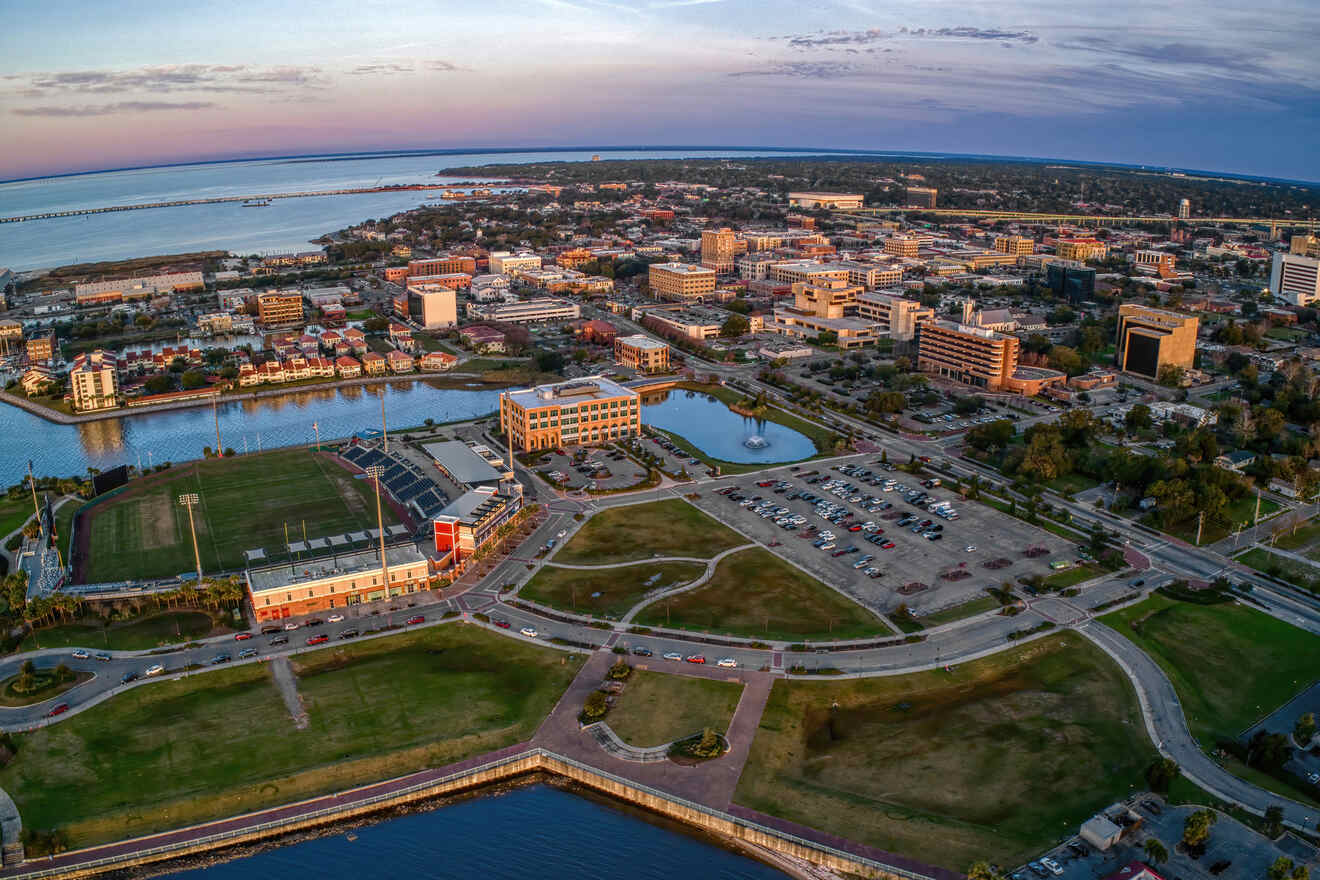 Aerial view of a coastal cityscape with buildings, a stadium, roads, and a waterway at sunset.