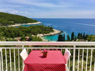 A table with a red tablecloth and a potted plant on a balcony overlooks a picturesque coastal landscape with trees, cliffs, and a body of water.