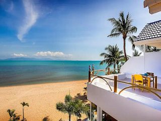 A beachfront balcony overlooks a sandy beach with palm trees and a clear blue ocean under a bright sky.
