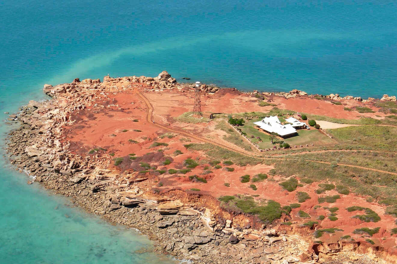 A rugged coastline with red rocks and a small white house on a peninsula surrounded by turquoise waters.
