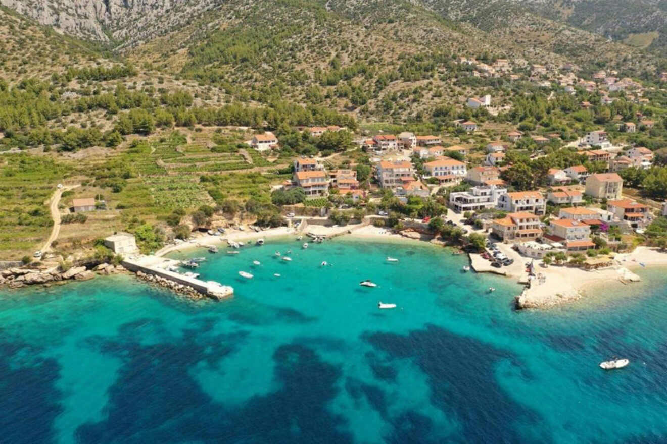 Aerial view of a coastal village with a small harbor, boats moored in turquoise water, and houses with red roofs surrounded by green hills.