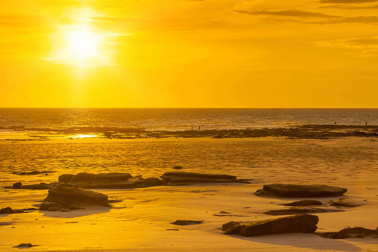 A beach during sunset, with golden light reflecting off the sand and rocks scattered across the shore.
