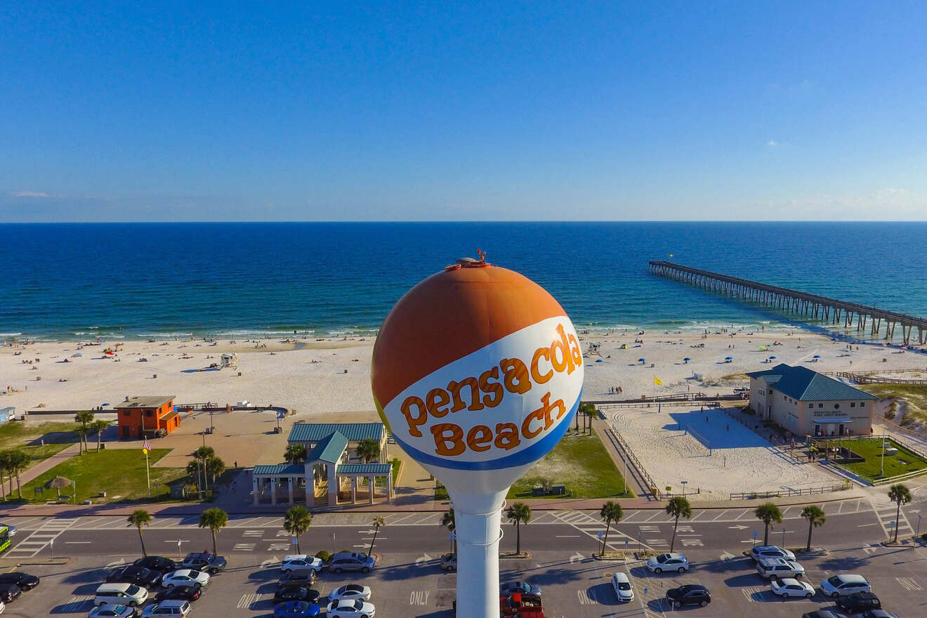 A water tower with "Pensacola Beach" written on it stands near a sandy beach and ocean, with a pier extending into the water. Cars are parked along the road in front.