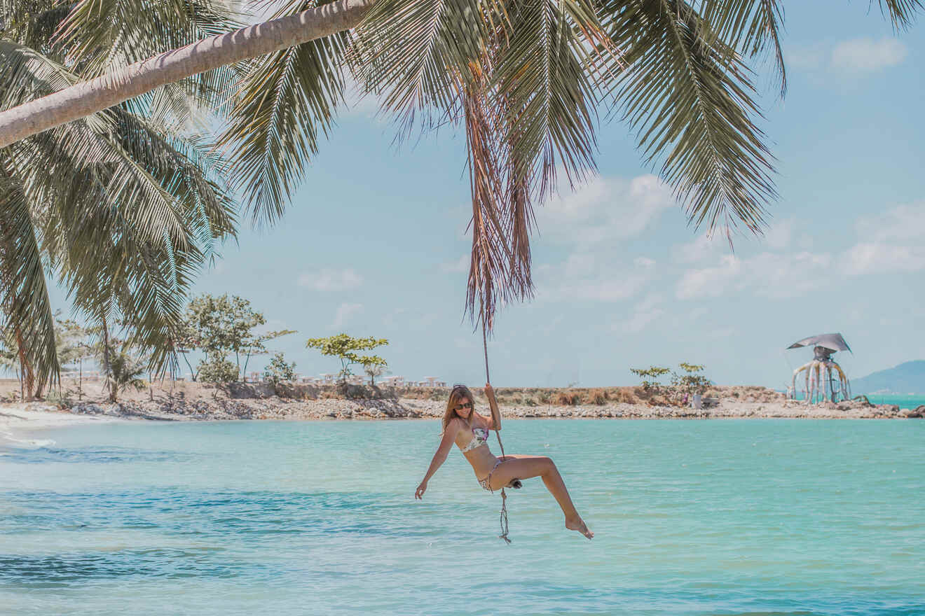 A person in a swimsuit swings from a palm tree over clear turquoise water at a tropical beach.