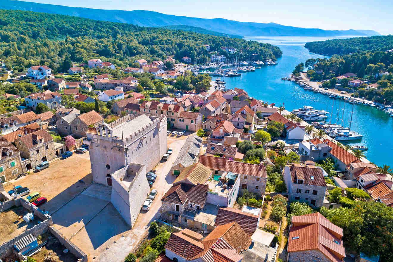 Aerial view of a historic coastal town with stone buildings, a prominent fortified structure, and a marina along a narrow waterway, surrounded by lush green forests and distant mountains.