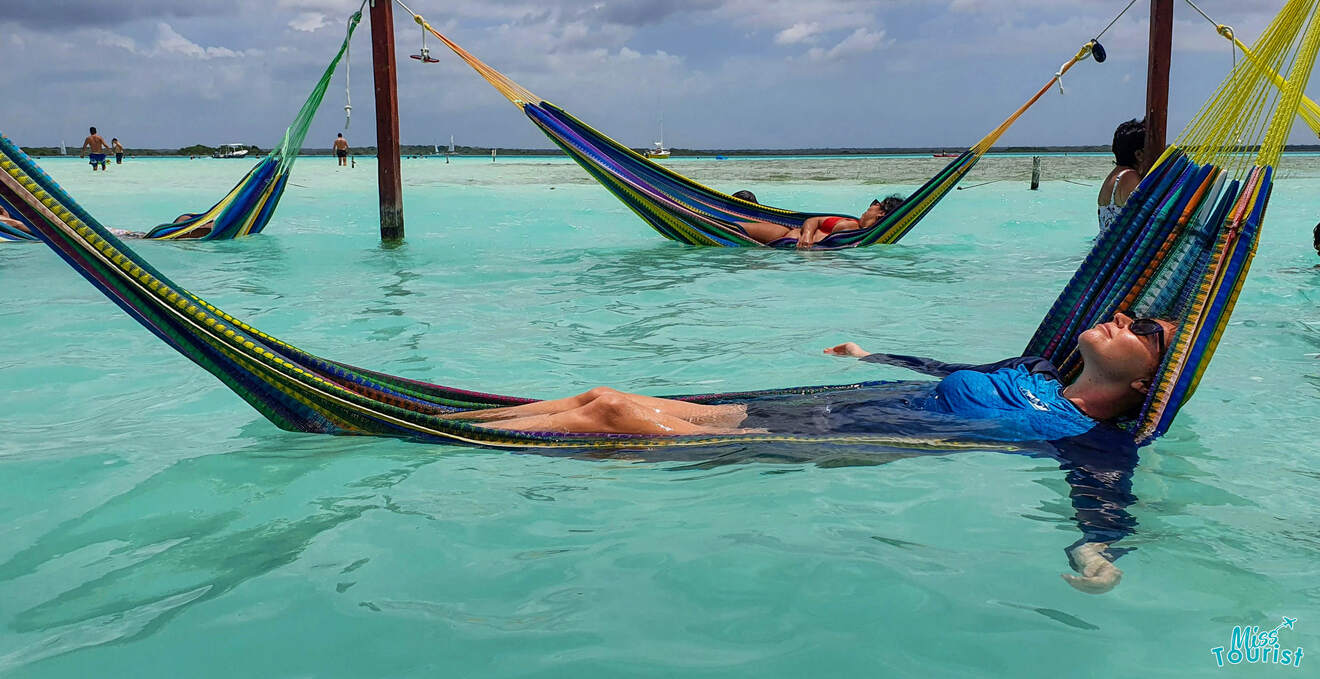 Person relaxing on a hammock suspended above clear turquoise water with other people in similar hammocks in the background.