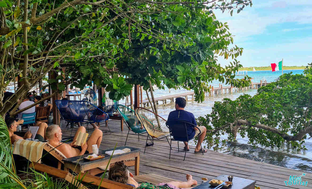 People relax on a wooden deck by the water's edge, with some sitting on chairs under large leafy trees and others lying in hammocks. A Mexican flag is visible across the water.