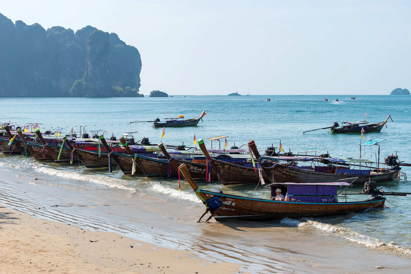 Serene beach scene with a lineup of colorful longtail boats docked on the shore, clear waters, and a picturesque view of limestone cliffs in the distance