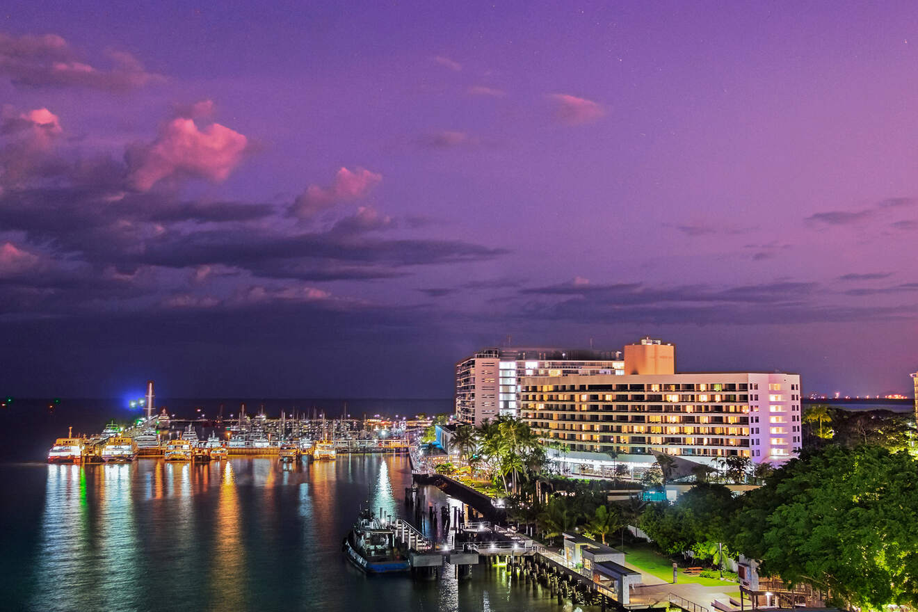 Nighttime view of a vibrant marina, illuminated buildings reflecting on the water with a dramatic purple sky above, possibly in a coastal city like Cairns, Australia