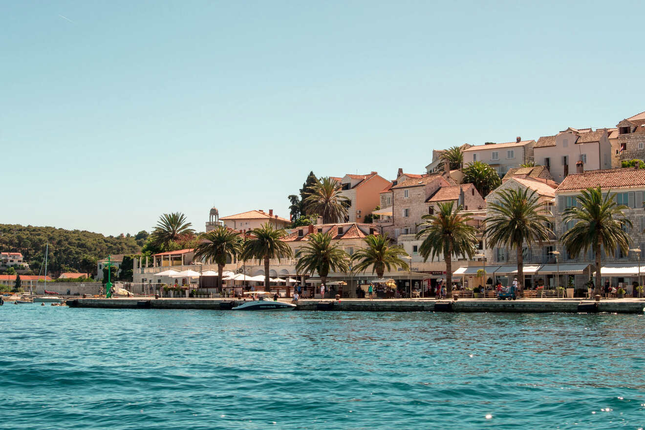 A seaside town with palm trees, waterfront buildings, and outdoor cafes. A boat is docked near the shore, and the water is calm and blue under a clear sky.