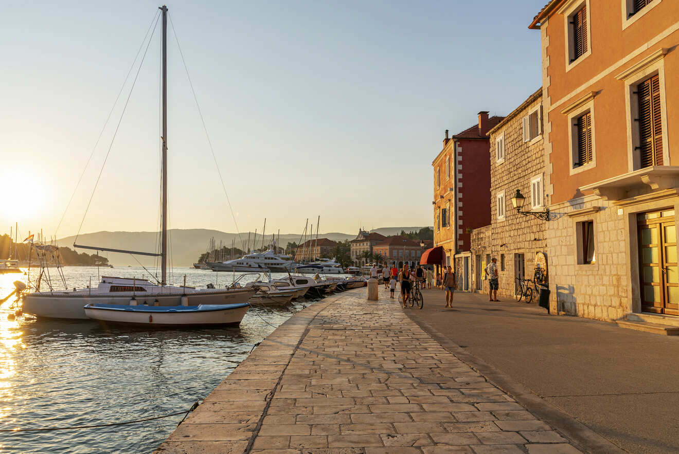 A seaside promenade at sunset with docked sailboats, people walking, and buildings lining the waterfront under a clear sky.