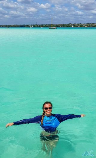 A woman wearing sunglasses and a blue long-sleeve shirt is standing waist-deep in clear turquoise water with boats and a distant shoreline visible in the background.