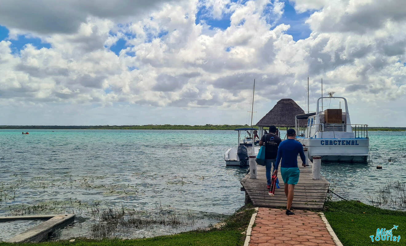 Three people walking on a wooden dock towards boats docked on a turquoise lake under a cloudy sky.