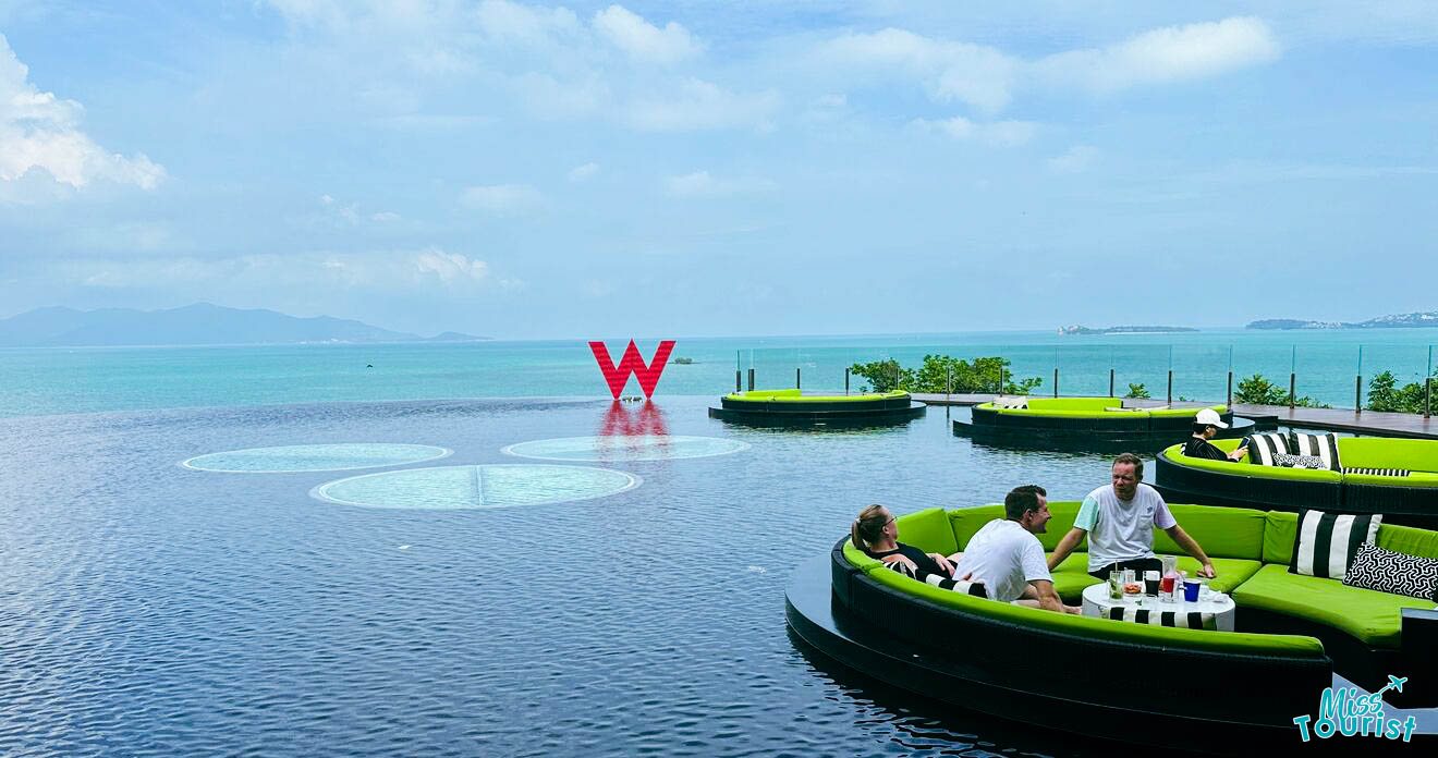 Three people sit on green outdoor seating beside an infinity pool overlooking the sea with a large red "W" sculpture in the background.