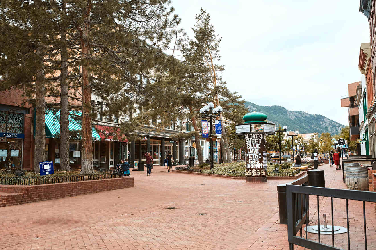 Pedestrian street with shops and trees, people walking, and mountains in the background, under a cloudy sky.