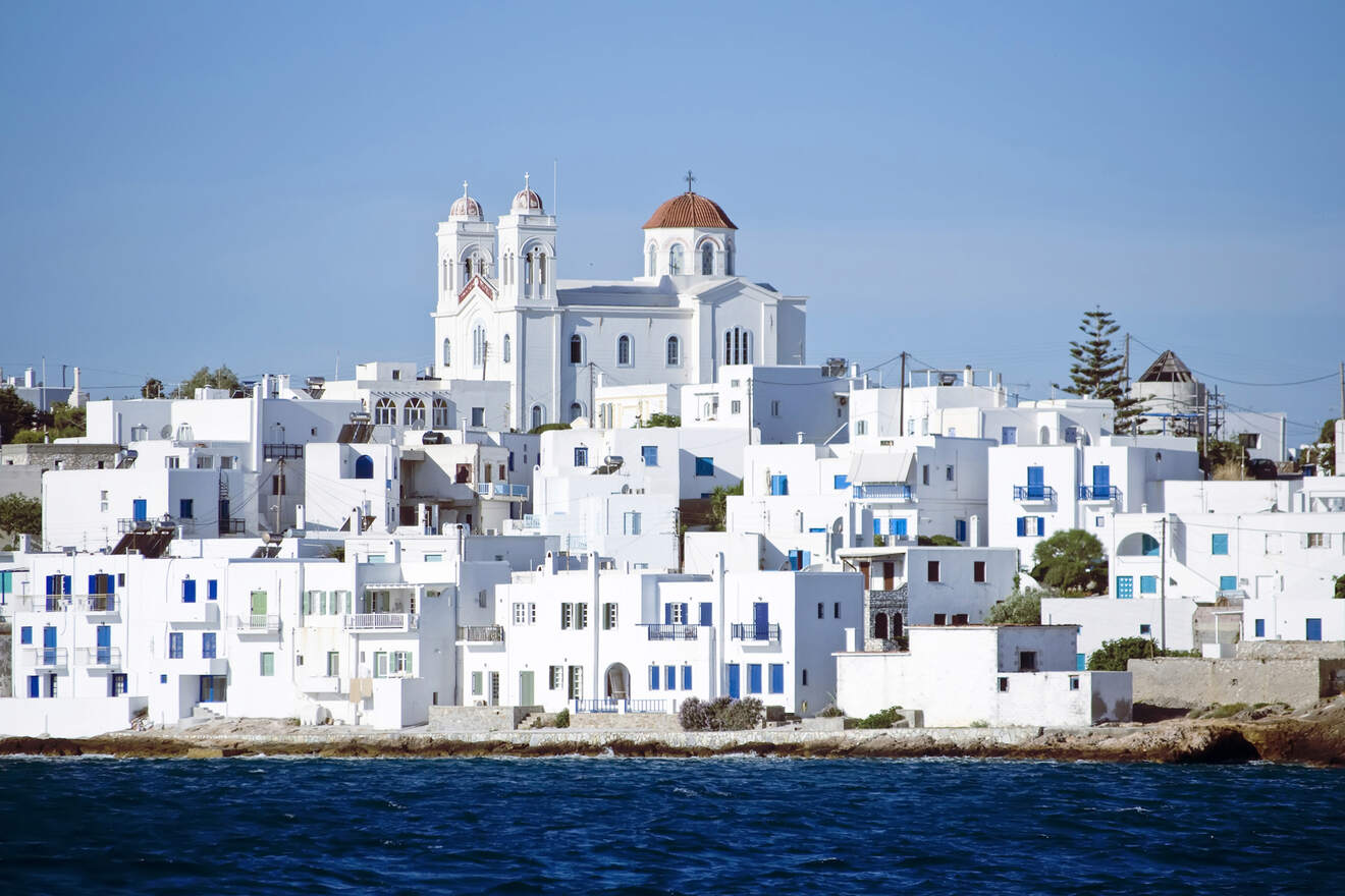 A coastal town with white buildings and a large church with domes is seen from the water, under a clear blue sky.