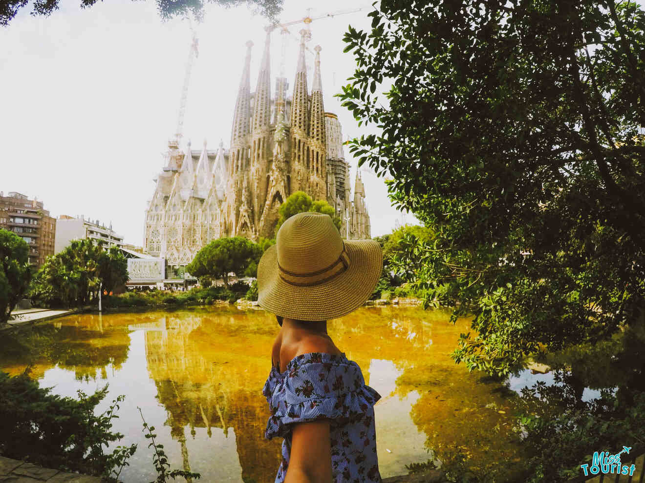 Yulia, the founder of this website,  in a hat and floral dress stands by a pond, looking at the Sagrada Familia, with construction cranes visible in the background.