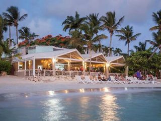 Beachfront dining area at dusk, illuminated by soft lights, with palm trees and a calm ocean in the background, creating a romantic ambiance