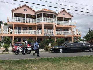 Modern three-story residential building in a suburban setting, with balconies on each level, captured during the daytime with pedestrians and vehicles passing by