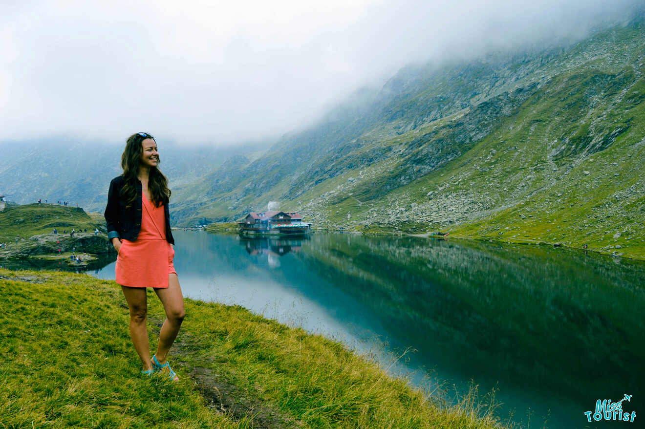 Yulia, the founder of this website, in a pink dress stands by a reflective lake surrounded by misty mountains.