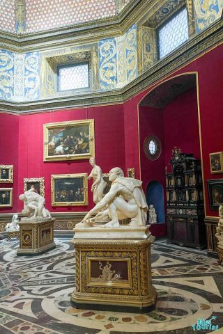 Statues and paintings in a richly decorated room with red walls, ornate ceiling, and tiled floor, featuring classical sculptures and framed artworks displayed prominently.