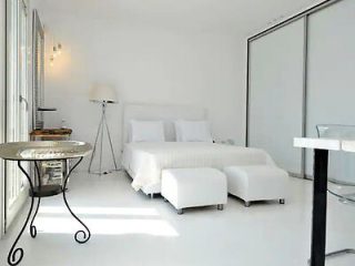 A minimalist white bedroom with a double bed, two ottomans at the foot, a round glass side table on the left, a floor lamp, and large sliding wardrobe doors.