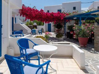 A sunny outdoor patio with blue chairs and a round table, surrounded by white buildings and vibrant red bougainvillea flowers overhead.