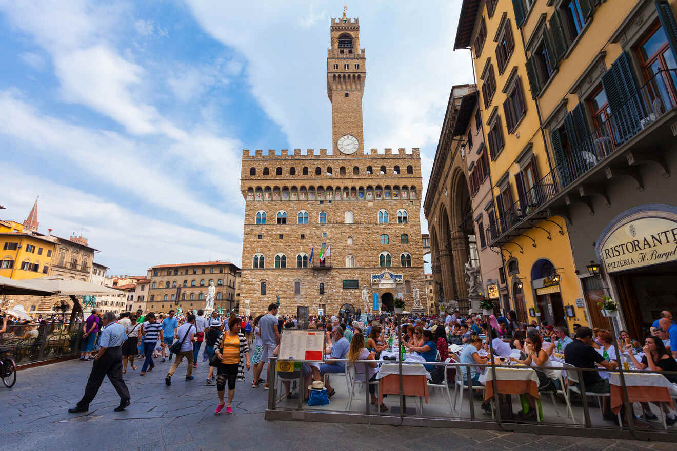 Tourists and locals gather in the bustling square in front of Palazzo Vecchio in Florence, Italy, with outdoor cafes and historic buildings surrounding the area.