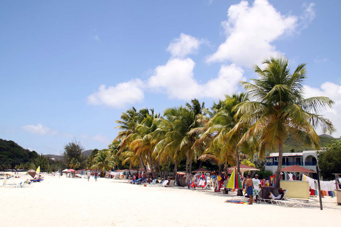 Sunny beach scene with visitors enjoying the white sand, lined with palm trees and colorful beach vendors under a clear blue sky.