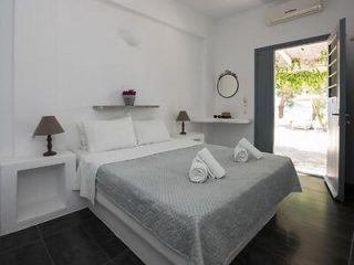 A minimalist bedroom with a double bed, white linens, grey blanket, two rolled towels, two bedside tables with lamps, a small round mirror, and an open door leading to a patio.