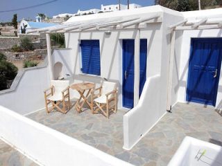 A white building with blue shutters and doors features a small terrace with a wooden table and two matching chairs on a stone-paved surface.