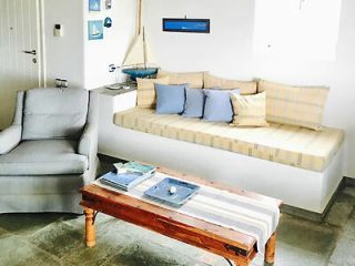 A small living room featuring a cushioned bench with plaid and solid pillows, a gray armchair, and a wooden coffee table with placemats and books. Nautical decor is on the walls.