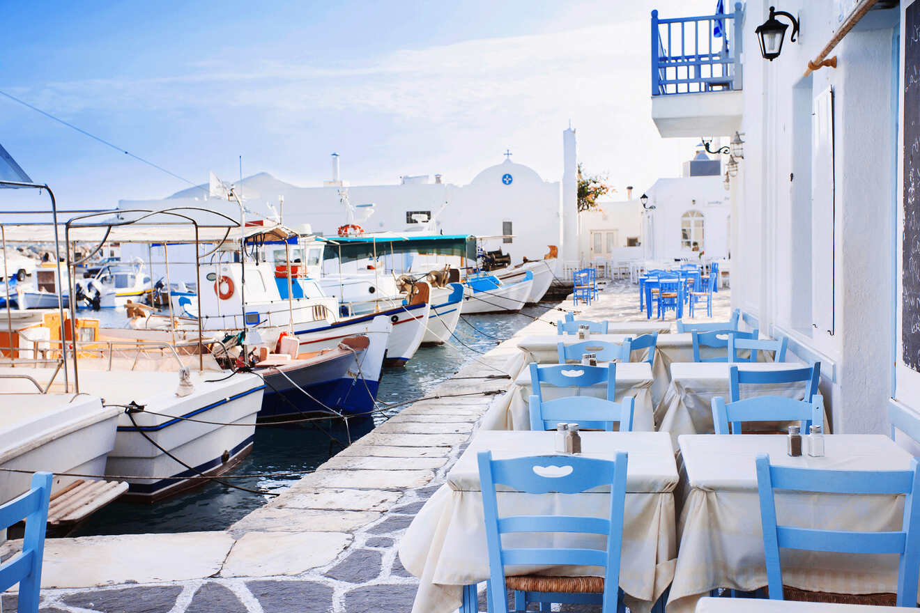 A serene coastal scene featuring an empty outdoor cafe with blue chairs and white tables. Fishing boats are docked along the waterfront, and white-washed buildings are seen in the background.