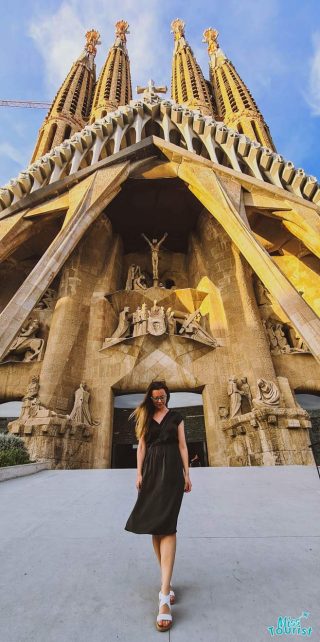 Yulia, the founder of this website,  in a black dress walks in front of the Sagrada Familia's intricate facade, with sunlight illuminating the towers.