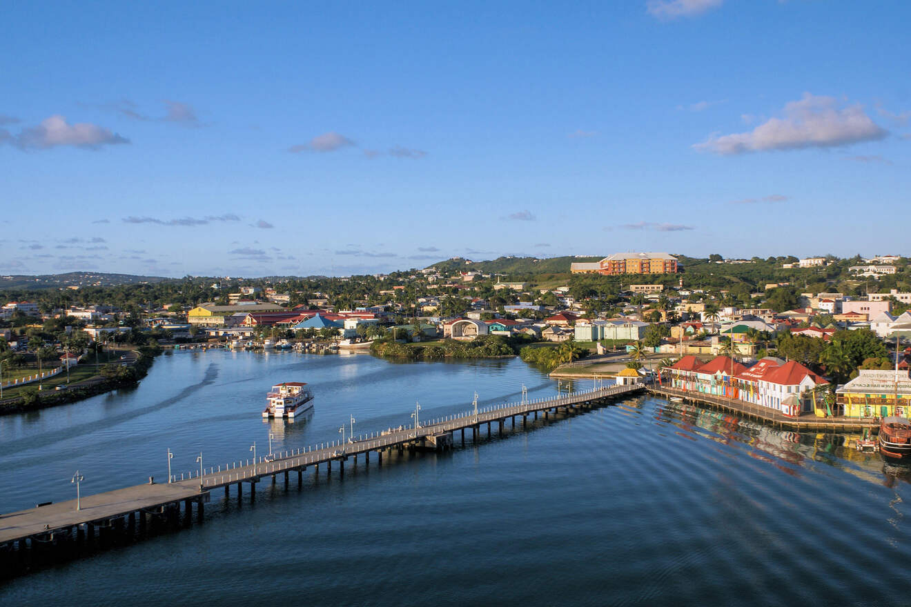 Aerial view of a coastal town with a long pier extending into the calm waters, surrounded by various buildings and lush green hills under a clear blue sky