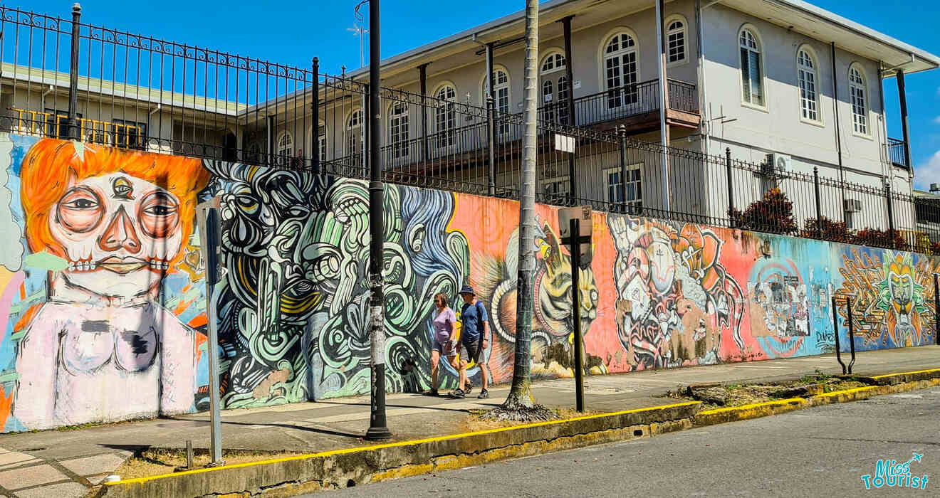 A colorful mural spans the length of a concrete wall along a sidewalk, featuring various artistic styles and figures. Two people walk past the mural in front of a large building with balconies.