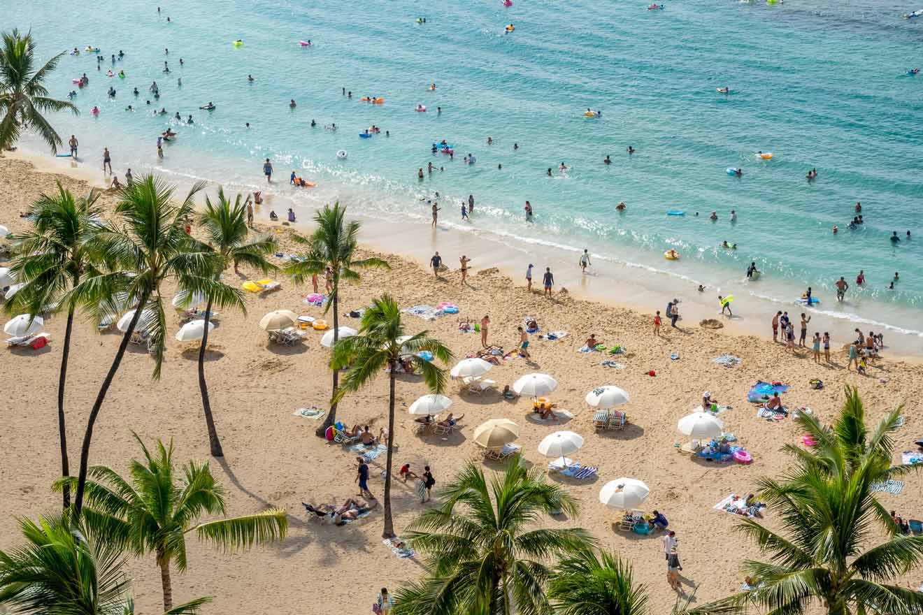 Aerial view of a sandy beach with numerous people swimming in the sea and sunbathing on the shore; palm trees and white umbrellas are scattered across the beach.