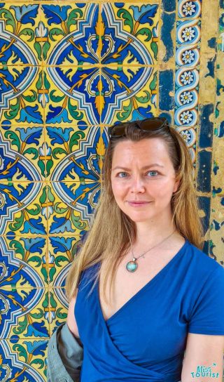 The writer of the post poses in front of a vibrant and intricate tiled wall, wearing a blue top and a necklace, smiling softly.