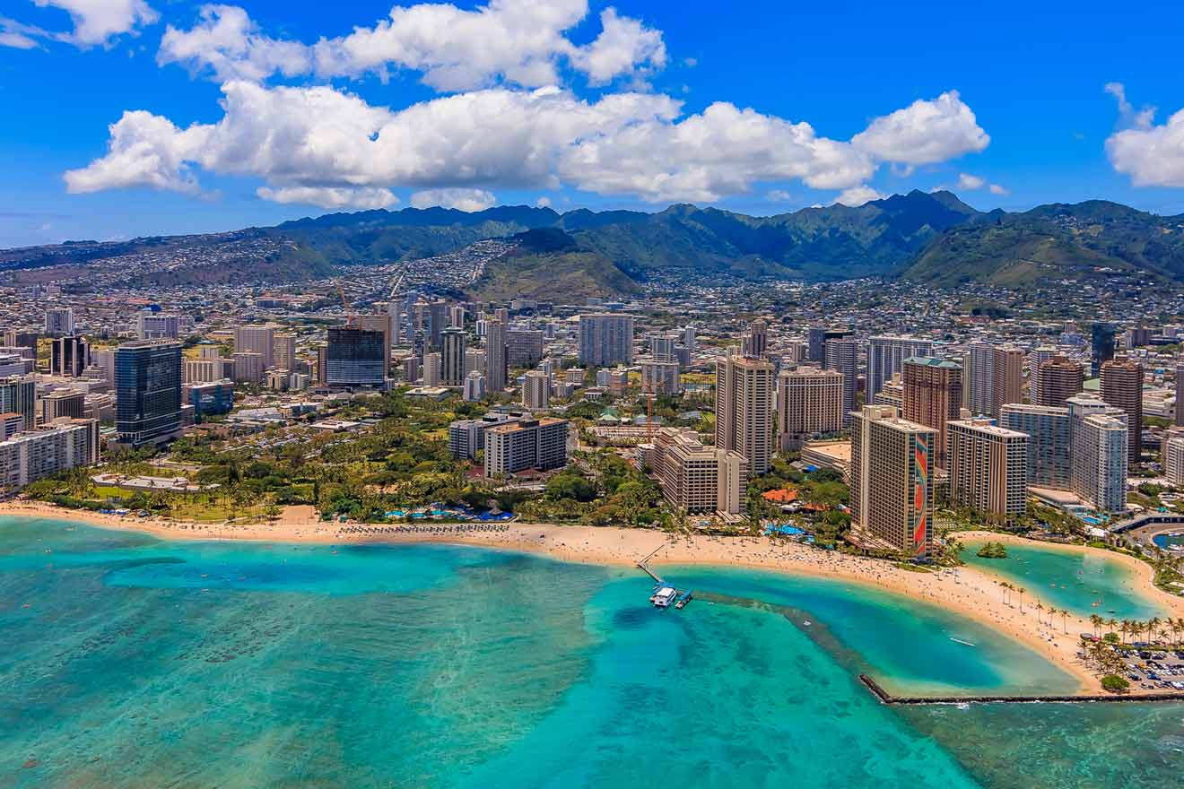 Aerial view of a coastal city with high-rise buildings, sandy beach, and turquoise waters, set against a backdrop of green mountains under a blue sky with scattered clouds.