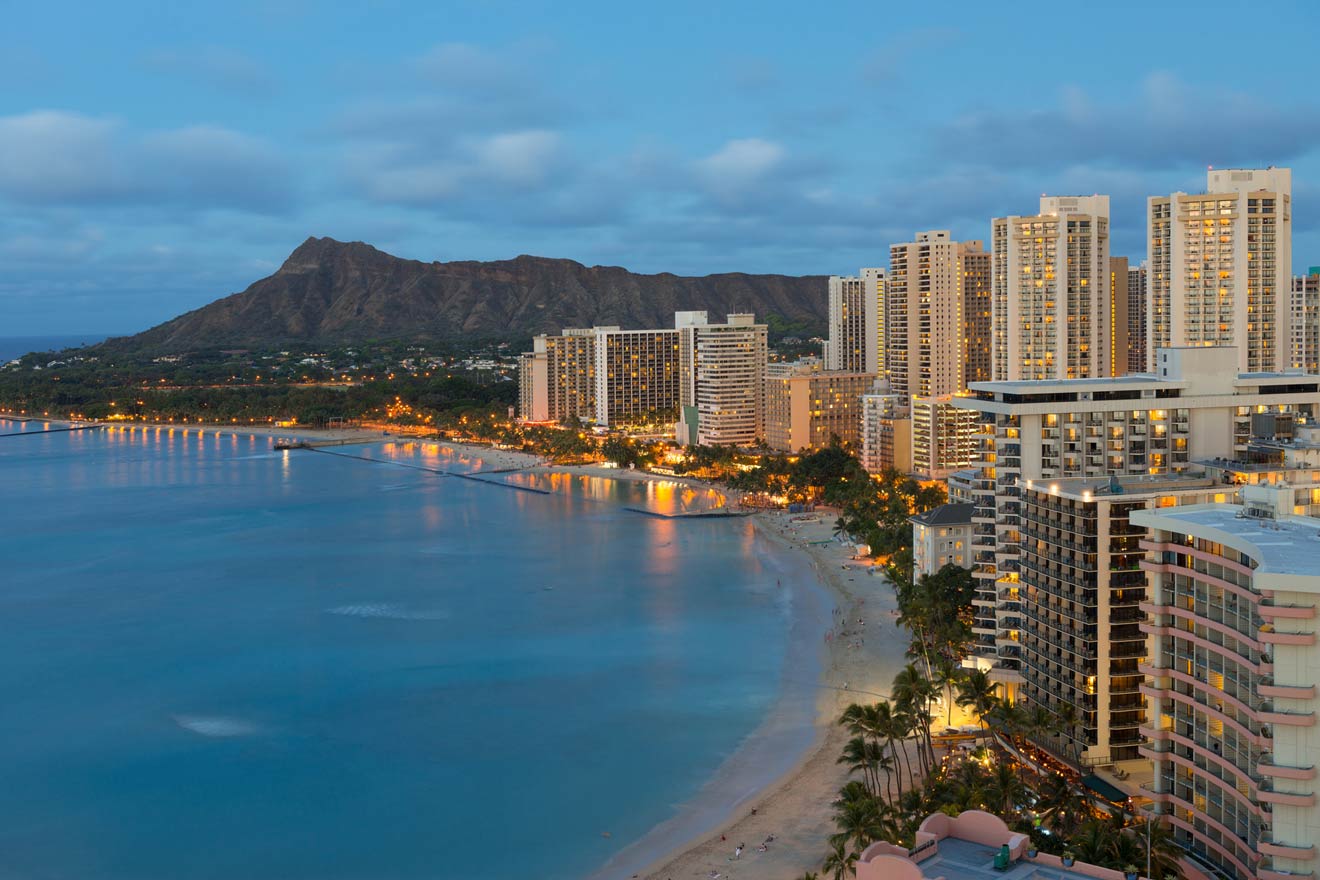 Evening view of a coastal cityscape with high-rise buildings, a sandy beach, and illuminated lights. A mountain range stands in the background under a cloudy sky.