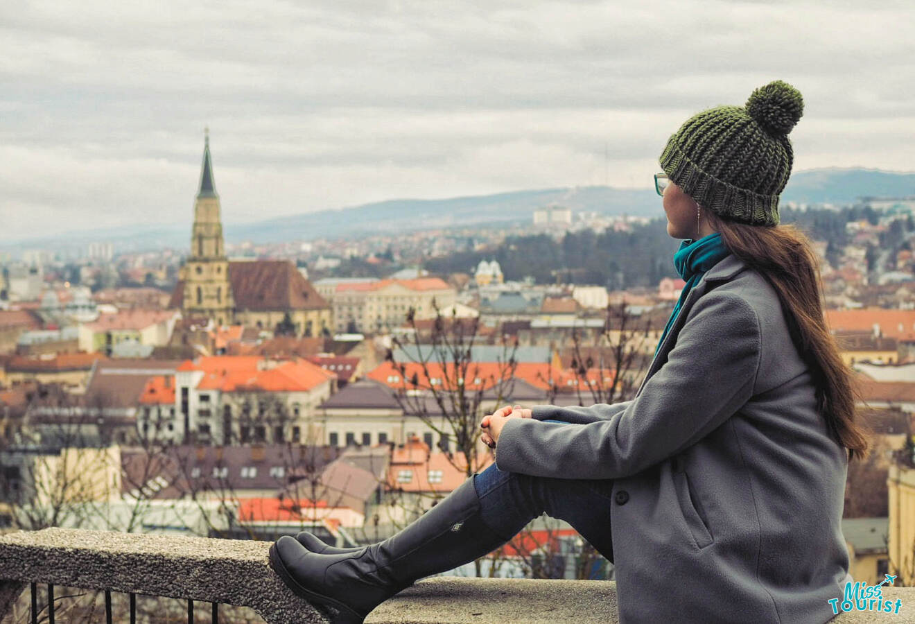 Yulia, the founder of this website, in a coat and beanie sits on a ledge, overlooking a cityscape with red-roofed buildings and a church tower in the distance.