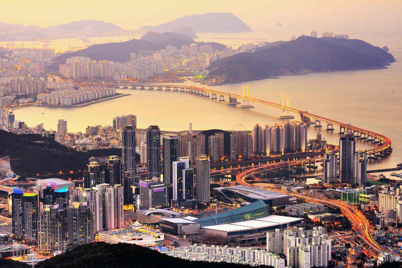 Aerial view of a coastal city with tall buildings, bridges, and mountains in the background at sunset.