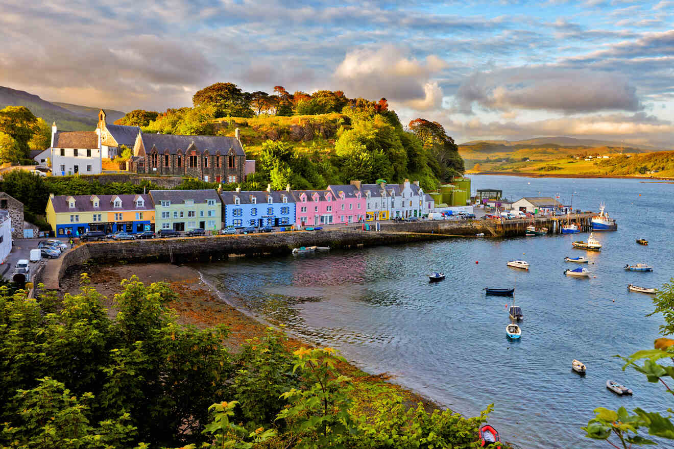 A picturesque seaside village with colorful buildings lining the waterfront, small boats docked in the harbor, and lush greenery on the hillside under a partly cloudy sky.