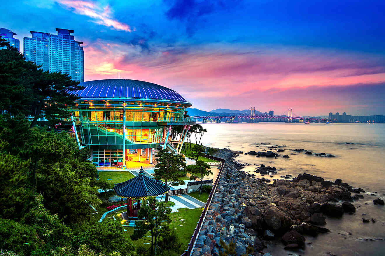 Modern building with colorful lighting by the coast, with a bridge and city skyline at sunset.