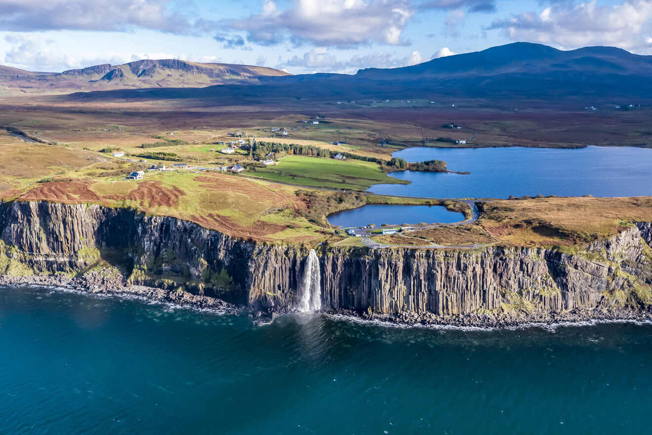 Aerial view of a coastal landscape with cliffs, a waterfall falling into the ocean, fields, and a lake surrounded by hills under a partly cloudy sky.