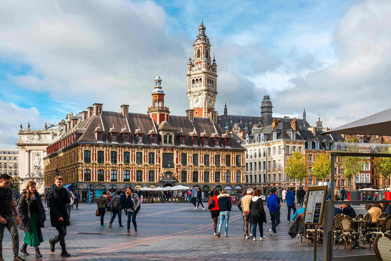 People walking and sitting at a café in a large open square with historic buildings and a tall clock tower in the background.