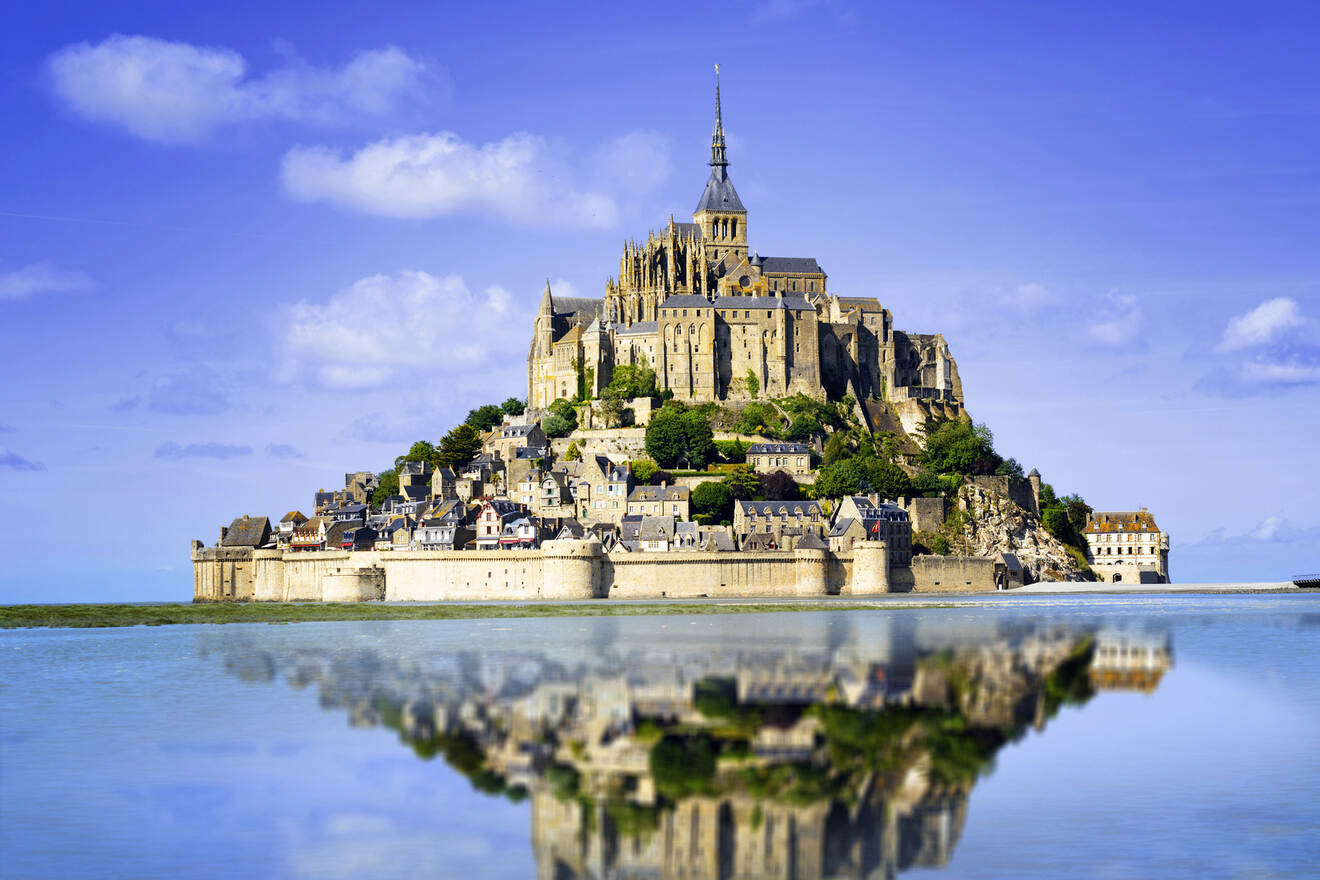 Mont Saint-Michel, a historic island commune in Normandy, France, surrounded by water at high tide and reflecting in the calm water below, with a clear blue sky in the background.